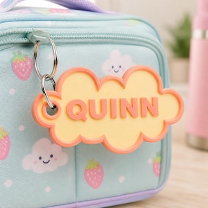 Pu&ograve; includere: Una borsa per il pranzo azzurra con cerniera, decorata con fragole e nuvole. Un'etichetta a forma di nuvola con il nome "QUINN" in lettere arancioni pende dalla cerniera. L'etichetta ha un centro giallo e un bordo arancione.