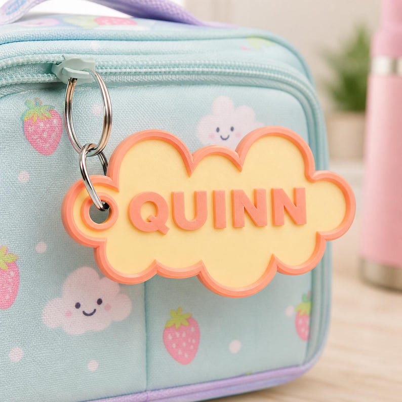 Pu&ograve; includere: Una borsa per il pranzo azzurra con cerniera, decorata con fragole e nuvole. Un'etichetta a forma di nuvola con il nome "QUINN" in lettere arancioni pende dalla cerniera. L'etichetta ha un centro giallo e un bordo arancione.