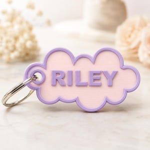 Pu&ograve; includere: Un portachiavi personalizzato a forma di nuvola, con il nome "RILEY" in lettere color lavanda. Il ciondolo a forma di nuvola ha un centro rosa chiaro e un bordo lavanda, attaccato a un portachiavi argentato.