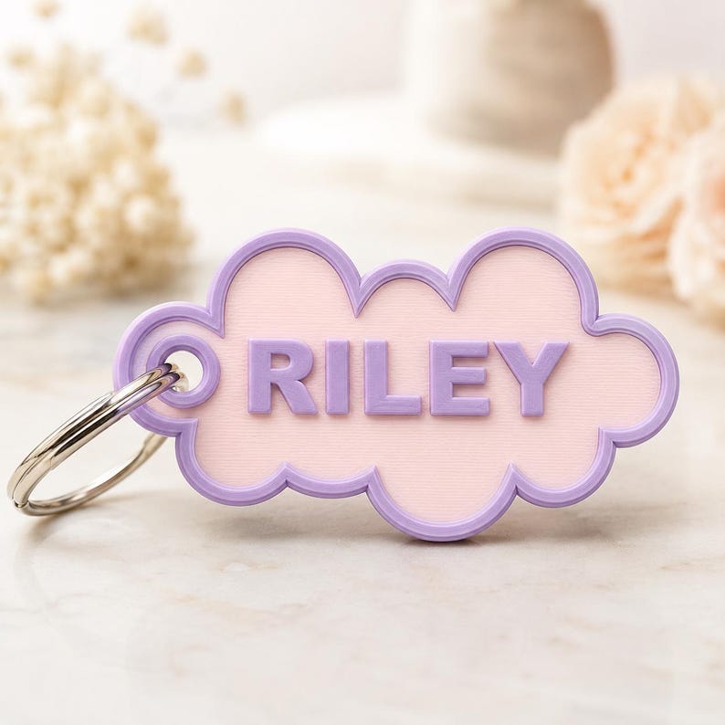 Pu&ograve; includere: Un portachiavi personalizzato a forma di nuvola, con il nome "RILEY" in lettere color lavanda. Il ciondolo a forma di nuvola ha un centro rosa chiaro e un bordo lavanda, attaccato a un portachiavi argentato.
