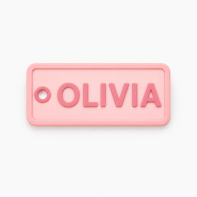 May include: A rectangular, light pink name tag with rounded corners. The name "OLIVIA" is displayed in raised, darker pink letters. A small, circular detail is on the left side of the tag.