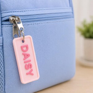 May include: Close-up of a light blue backpack with a silver zipper and a pink rectangular tag. The tag reads "DAISY" in raised pink letters. A small mesh pocket is visible on the side of the backpack.