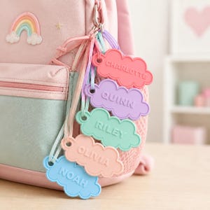 Pu&ograve; includere: Uno zaino rosa con una toppa a forma di arcobaleno e una collezione di targhette con nomi a forma di nuvola. I tag sono in vari colori pastello, ognuno con un nome diverso: Charlotte, Quinn, Riley, Olivia e Noah. I tag sono attaccati allo zaino con un fermaglio di metallo.