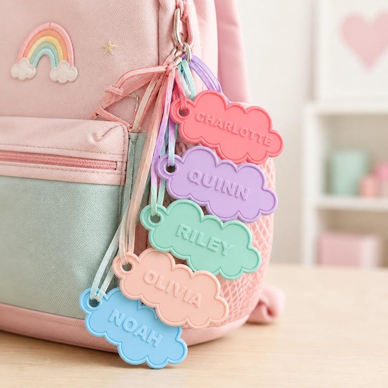 Pu&ograve; includere: Uno zaino rosa con una toppa a forma di arcobaleno e una collezione di targhette con nomi a forma di nuvola. I tag sono in vari colori pastello, ognuno con un nome diverso: Charlotte, Quinn, Riley, Olivia e Noah. I tag sono attaccati allo zaino con un fermaglio di metallo.