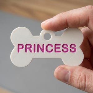 May include: A white bone-shaped pet identification tag featuring the word PRINCESS embossed in bright pink capital letters. The tag has a circular hole at the top for attachment to a collar, with a smooth, matte finish and clean, defined edges.