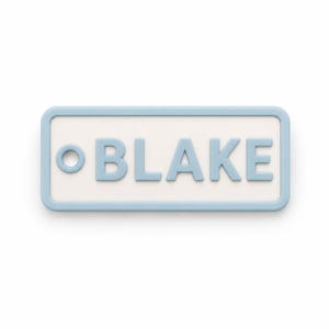 May include: A rectangular name tag with rounded corners. The tag has a light blue border and the name "BLAKE" in light blue, raised letters. The background is white. The tag is a simple, clean design.