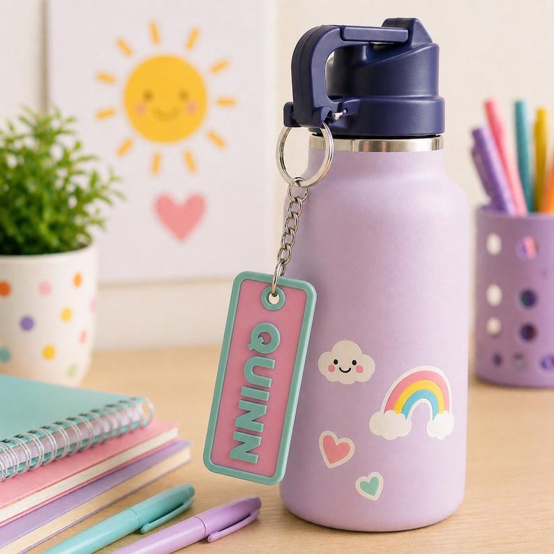 May include: A light purple water bottle with a dark blue lid and handle, adorned with a rainbow, cloud, and heart stickers. A keychain with the name "Quinn" hangs from the bottle. Pens and notebooks are in the background.