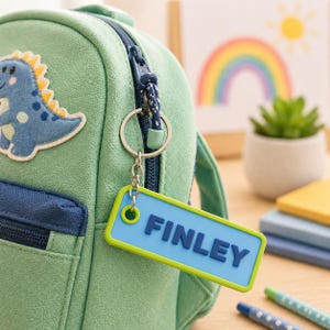 May include: A green backpack with a blue dinosaur patch and a blue and green keychain tag that reads "FINLEY". The tag is attached to the zipper with a silver keyring. A rainbow and sun illustration is in the background.