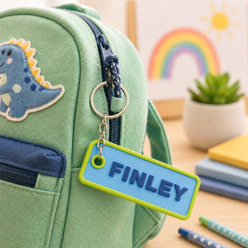 May include: A green backpack with a blue dinosaur patch and a blue and green keychain tag that reads "FINLEY". The tag is attached to the zipper with a silver keyring. A rainbow and sun illustration is in the background.
