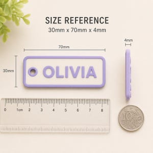 May include: A lavender and white rectangular keychain with the name "OLIVIA" in raised letters. The keychain measures 30mm x 70mm x 4mm. A ruler and a coin are also in the image.