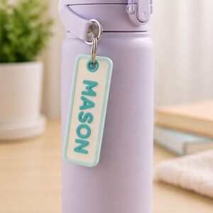 May include: A light purple insulated water bottle with a matching lid and handle. A small, rectangular tag with the name "MASON" in teal letters hangs from the bottle. The bottle is designed to keep drinks cold or hot.
