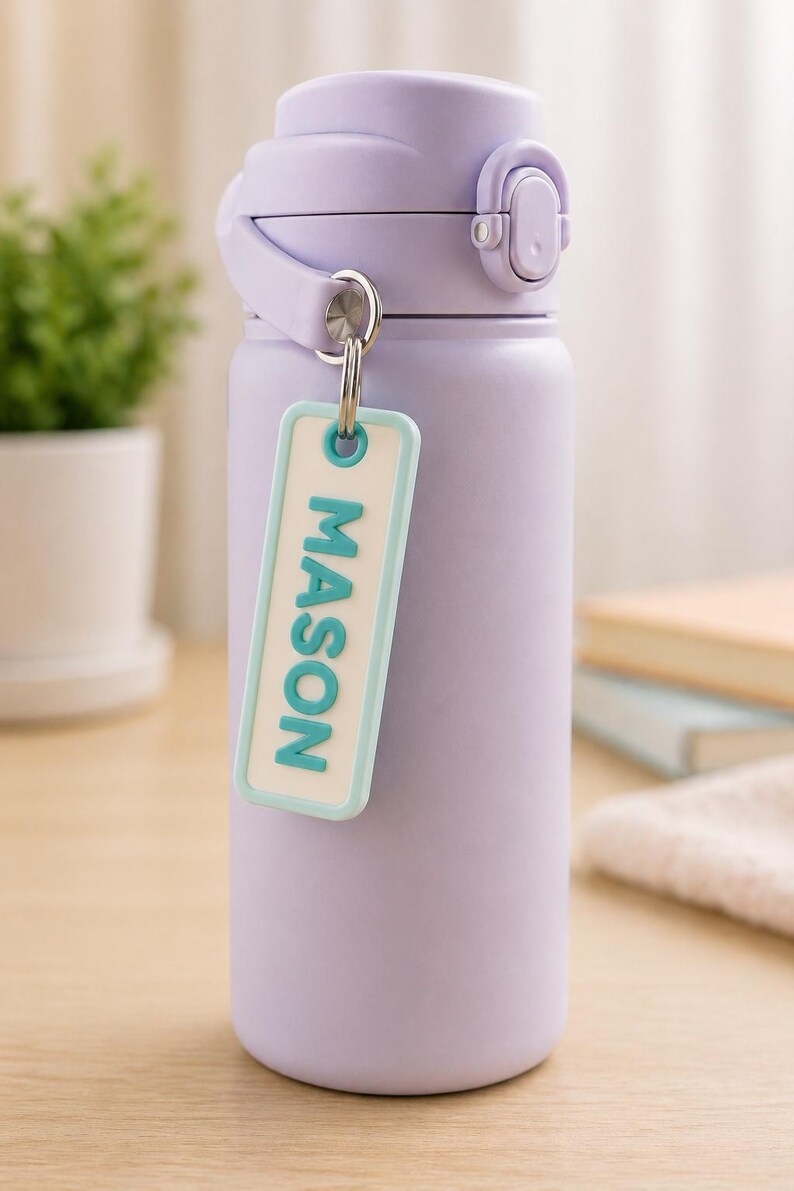 May include: A light purple insulated water bottle with a matching lid and handle. A small, rectangular tag with the name "MASON" in teal letters hangs from the bottle. The bottle is designed to keep drinks cold or hot.