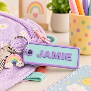 May include: A light blue rectangular keychain with the name "JAMIE" in purple letters. The keychain is attached to a keyring with keys and a rainbow charm. A purple daisy-patterned bag is in the background.