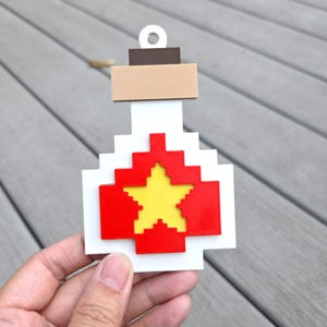 Peut inclure: Un ornement en forme de bouteille pixelisée avec une base blanche, un centre en forme de croix rouge et une étoile jaune. Le haut comprend une section rectangulaire beige et un bouchon marron foncé avec un trou circulaire pour suspendre.
