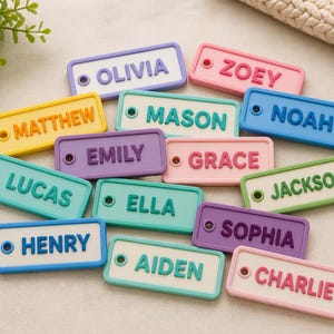 May include: Colorful name tags in various pastel colors, each with a different name in bold, raised letters. The tags are rectangular with rounded corners and a small hole on one side. Names include Olivia, Zoey, Matthew, Lucas, Mason, Emily, Grace, Noah, Jackson, Ella, Sophia, Henry, Aiden, and Charlie.