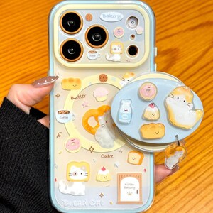 May include: A light blue and yellow phone case with a bakery theme. The case features cartoon images of bread, donuts, and cats. The text "Bakery", "Butter", "Cake", "Bread Cat", and "Kitten Bakery" are visible. The case also has a charm with a cat.