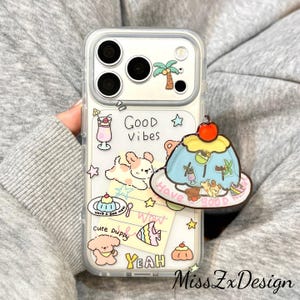 May include: A clear phone case featuring colorful cartoon illustrations of a puppy, a fish, a palm tree, and a drink. The text Good Vibes, Cute Puppy, and Yeah are printed on the back. A large, raised jelly dessert charm with a cherry on top is attached.