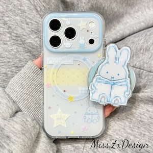May include: A clear smartphone case with a light blue camera frame, featuring Miffy character graphics, yellow and blue stars, and a circular magnetic ring. A matching Miffy-shaped phone grip with a blue dotted pattern is attached to the back.