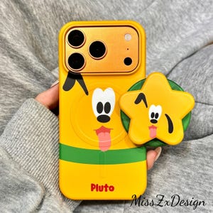 Puede incluir: Una funda de smartphone amarilla con un gráfico del personaje Pluto con un collar verde y el nombre Pluto impreso en rojo. En la parte posterior hay un soporte para teléfono en forma de estrella con la cara de un Pluto guiñando un ojo.
