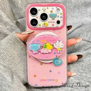 May include: A pink smartphone case featuring a grid pattern, colorful stars, and cartoon animal graphics. The case includes a circular pop-out grip with the text Be Good Friends and a small blue heart charm. The camera area is outlined in pink.