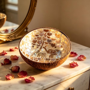 May include: A decorative bowl crafted from a coconut shell, showcasing a mosaic interior with an elephant and tree motif. The bowl is brown with a mosaic interior of white, gold, and brown tiles. Dried rose petals are scattered around the bowl.