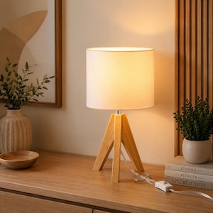 May include: A table lamp with a cylindrical white fabric shade and a tripod base made of light-colored wood. The lamp features a clear power cord with an inline rocker switch, emitting a warm, soft glow from the light source within the shade.