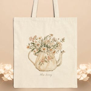 May include: A cream-colored canvas tote bag featuring a printed illustration of a ceramic teapot filled with a bouquet of white and light pink wildflowers. The text "Slow Living" is printed in a delicate, cursive font below the teapot.