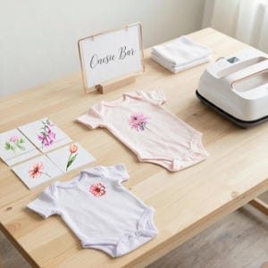 May include: A light pink baby bodysuit with a pink flower graphic and a white baby bodysuit with a pink flower graphic. A sign reads Onesie Bar. Four square cards feature watercolor flower illustrations. A white heat press machine sits nearby.