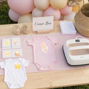May include: A Onesie Bar setup featuring a pink baby bodysuit with a popsicle graphic, a white baby bodysuit with a sun graphic, a white heat press, a sign reading Onesie Bar, small jars of wooden clips, and four cards with various cute graphics.