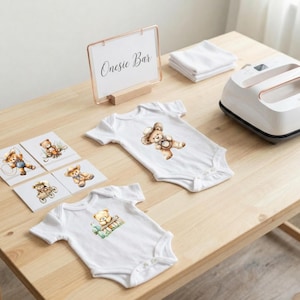 May include: Two white baby bodysuits featuring watercolor teddy bear illustrations, a sign reading Onesie Bar, a stack of white fabric squares, and a white heat press machine. The bears are depicted in various poses, including wearing hats.