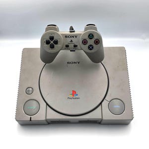 May include: A vintage Sony PlayStation console with a matching controller. The console is light gray with a circular disc tray and the PlayStation logo. The controller is also light gray with directional buttons and action buttons. The word "SONY" is visible on both.