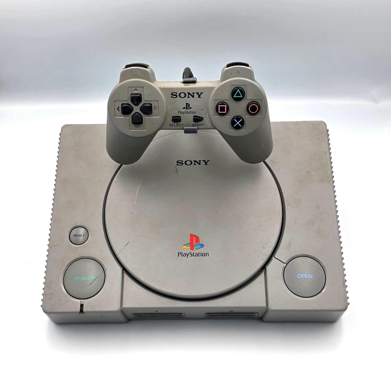 May include: A vintage Sony PlayStation console with a matching controller. The console is light gray with a circular disc tray and the PlayStation logo. The controller is also light gray with directional buttons and action buttons. The word "SONY" is visible on both.