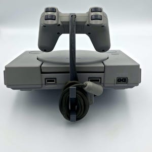 May include: A gray PlayStation console with a matching controller. The controller is positioned above the console, connected by a black cable. The console has a disc tray and various ports. The overall aesthetic is retro and minimalist.