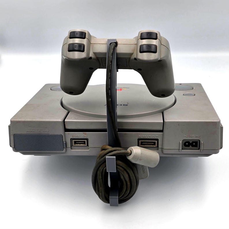May include: A gray PlayStation 1 console with a matching controller. The controller is connected to the console with a black cable. The console has several ports and a disc tray. The PlayStation 1 is a classic video game console.