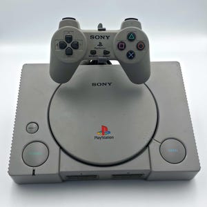 Puede incluir: Una consola PlayStation gris de Sony con un mando a juego. La consola tiene una bandeja de disco circular y el logotipo de PlayStation. El mando tiene botones direccionales, botones de acción y el logotipo de Sony.
