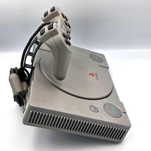 May include: A gray PlayStation console with a matching controller. The console has a disc tray, power and reset buttons, and the PlayStation logo. The controller is connected to the console with a black cable.