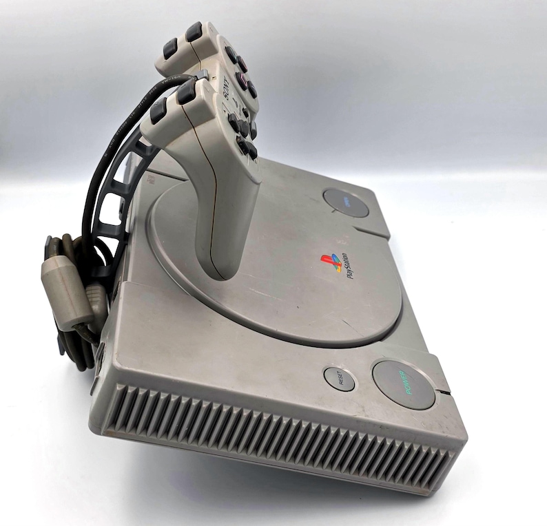 May include: A gray PlayStation console with a matching controller. The console has a disc tray, power and reset buttons, and the PlayStation logo. The controller is connected to the console with a black cable.