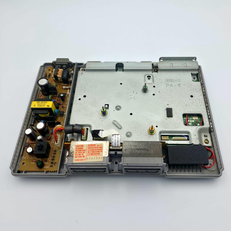 May include: An open, gray PlayStation 1 console interior, showcasing the internal components. The image reveals circuit boards, wiring, and a metal shield. A red and white caution label is visible.