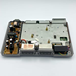 May include: An open, gray PlayStation console, revealing internal components. The top cover is off, showing circuit boards, connectors, and a metal plate. The console's ports and power supply are visible, with a "CAUTION" label.