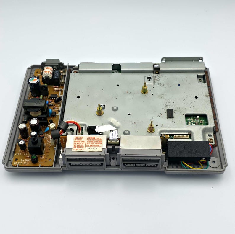 May include: An open, gray PlayStation console, revealing internal components. The top cover is off, showing circuit boards, connectors, and a metal plate. The console's ports and power supply are visible, with a "CAUTION" label.