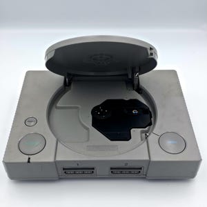 May include: A gray PlayStation 1 console with the disc tray open. The console has a power button, a reset button, and an open button. The disc tray is open, revealing the disc drive. The console has two controller ports.