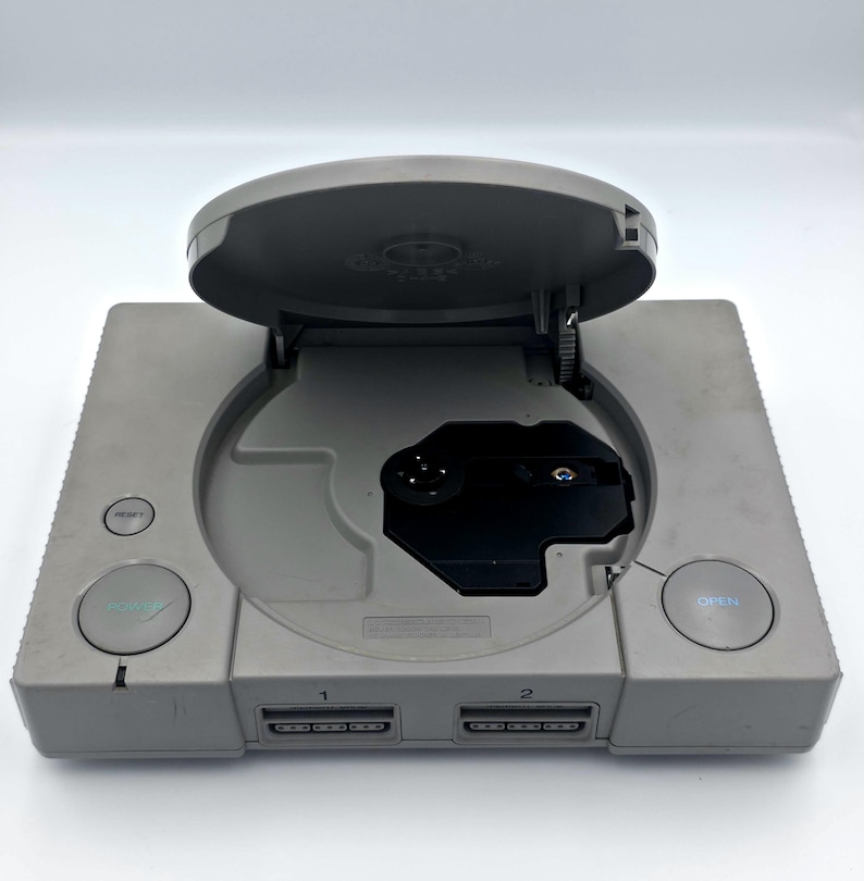 May include: A gray PlayStation 1 console with the disc tray open. The console has a power button, a reset button, and an open button. The disc tray is open, revealing the disc drive. The console has two controller ports.