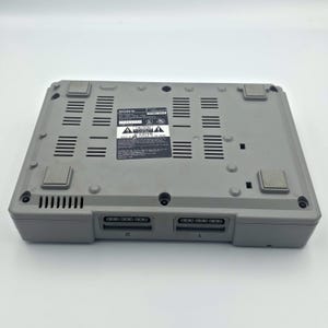 May include: A gray Sony PlayStation console, viewed from the bottom. The console has a rectangular shape with multiple vents and two controller ports. A label with text is visible.