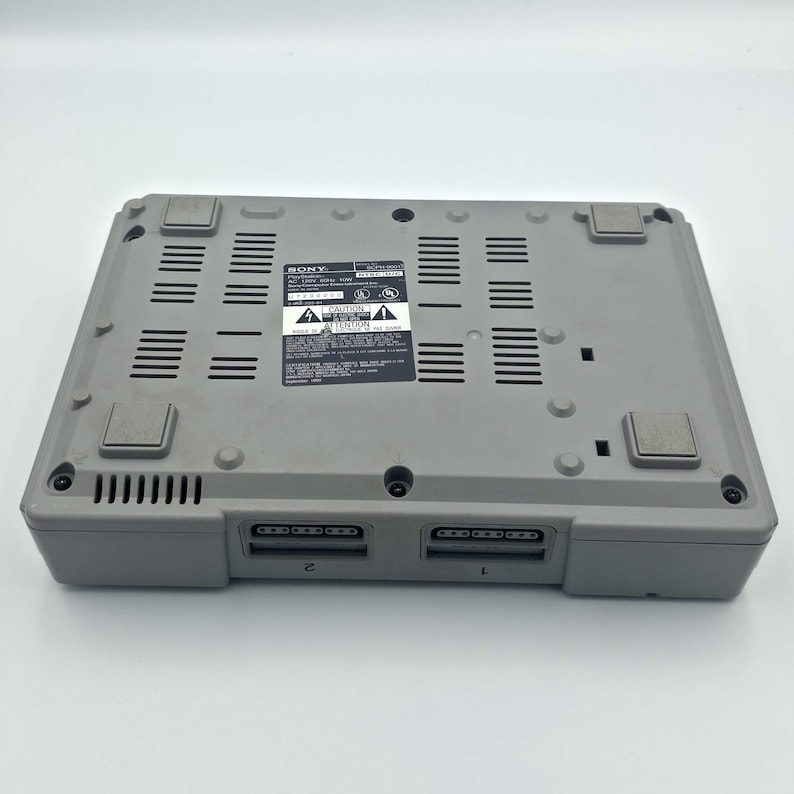 May include: A gray Sony PlayStation console, viewed from the bottom. The console has a rectangular shape with multiple vents and two controller ports. A label with text is visible.