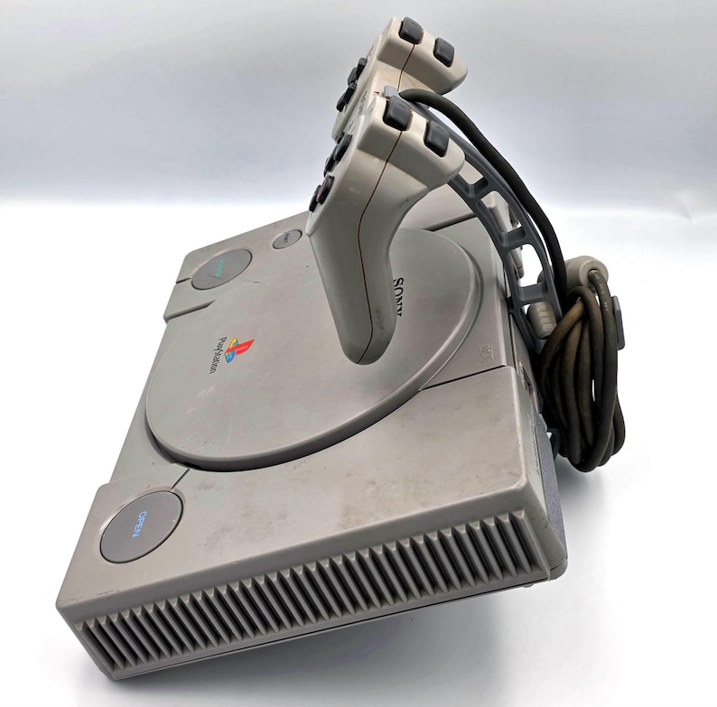 May include: A gray PlayStation console with two attached controllers. The console has a disc tray and several buttons. The controllers are gray with black buttons and a curved design. The PlayStation logo is visible.