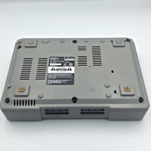 May include: A gray Sony PlayStation console, viewed from the bottom. The console has a rectangular shape with ventilation slots and two controller ports. A label with text and warning symbols is visible. The console is a classic gaming device.