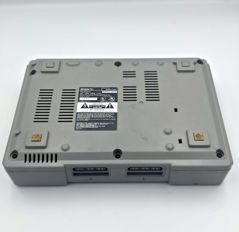 May include: A gray Sony PlayStation console, viewed from the bottom. The console has a rectangular shape with ventilation slots and two controller ports. A label with text and warning symbols is visible. The console is a classic gaming device.