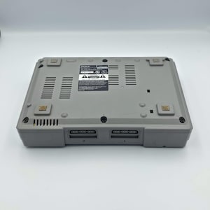 May include: A gray Sony PlayStation 1 game console, viewed from the bottom. The console has two controller ports labeled 1 and 2. The bottom features ventilation slots and several small, square rubber feet.