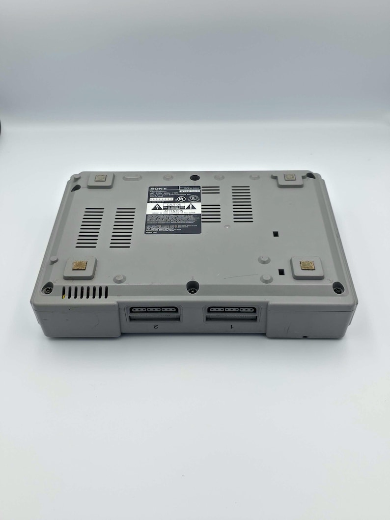 May include: A gray Sony PlayStation 1 game console, viewed from the bottom. The console has two controller ports labeled 1 and 2. The bottom features ventilation slots and several small, square rubber feet.