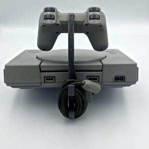 May include: A gray PlayStation console with a matching controller. The controller is connected to the console with a black cable. The console has a disc tray and several ports. The controller has a classic design with directional buttons and four action buttons.